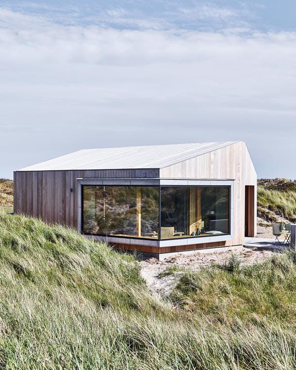 Het zomerhuis is bekleed met cederhout. De goten zijn verborgen onder het dak, waardoor de gevel en het dak gestroomlijnd in elkaar overlopen. Het zomerhuis is bekleed met cederhout. De goten zijn verborgen onder het dak, waardoor de gevel en het dak gestroomlijnd in elkaar overlopen.