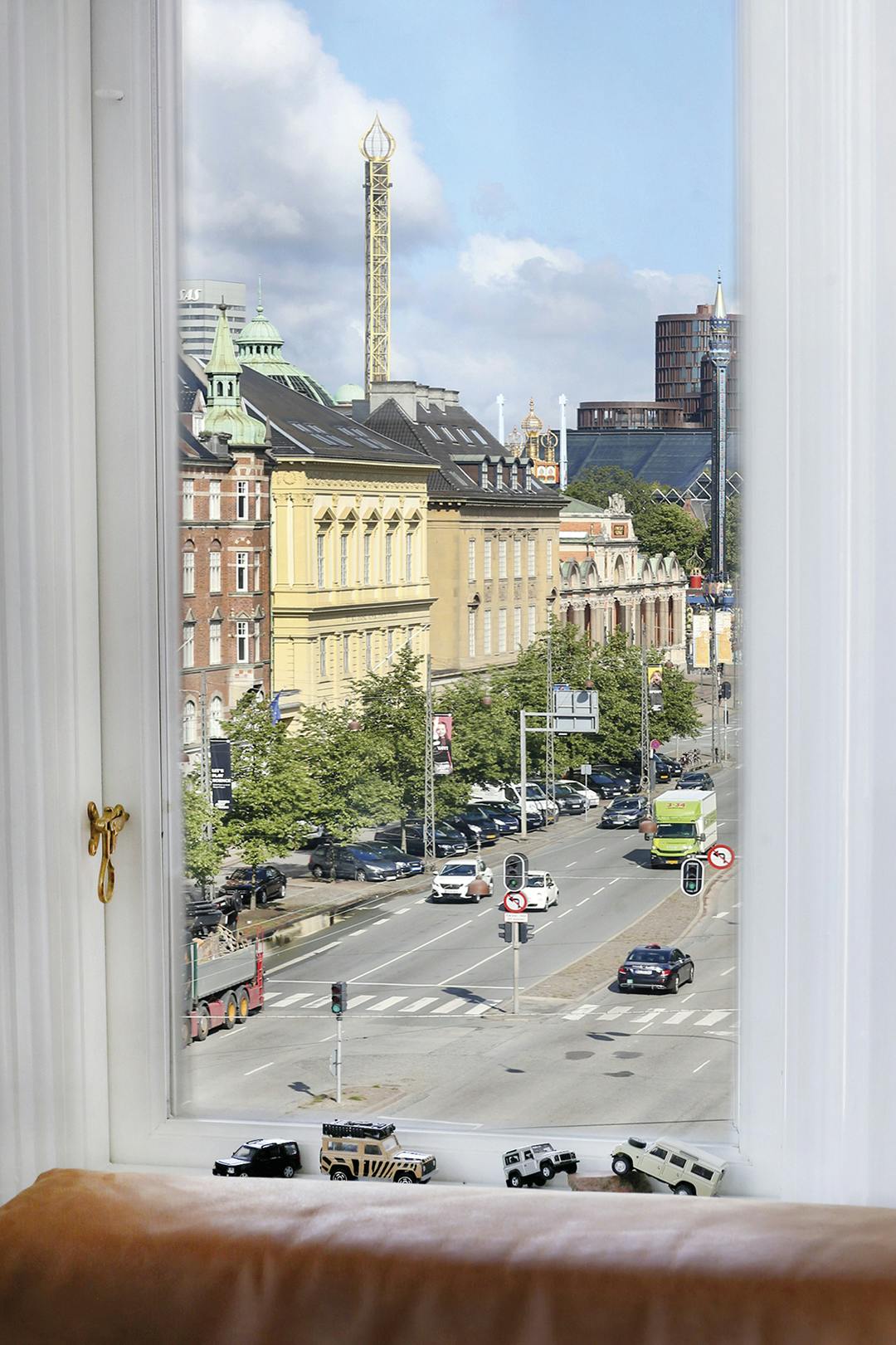 De vergulde toren van pretpark Tivoli is vanuit de woonkamer te zien. Op de vensterbank staat een collectie Land Rovers, die de bewoners herinnert aan de Land Rovers die ze zelf hadden voor ze overgingen op een elektrische auto. Uitzicht op Tivoli Kopenhagen, Land Rovers in vensterbank.