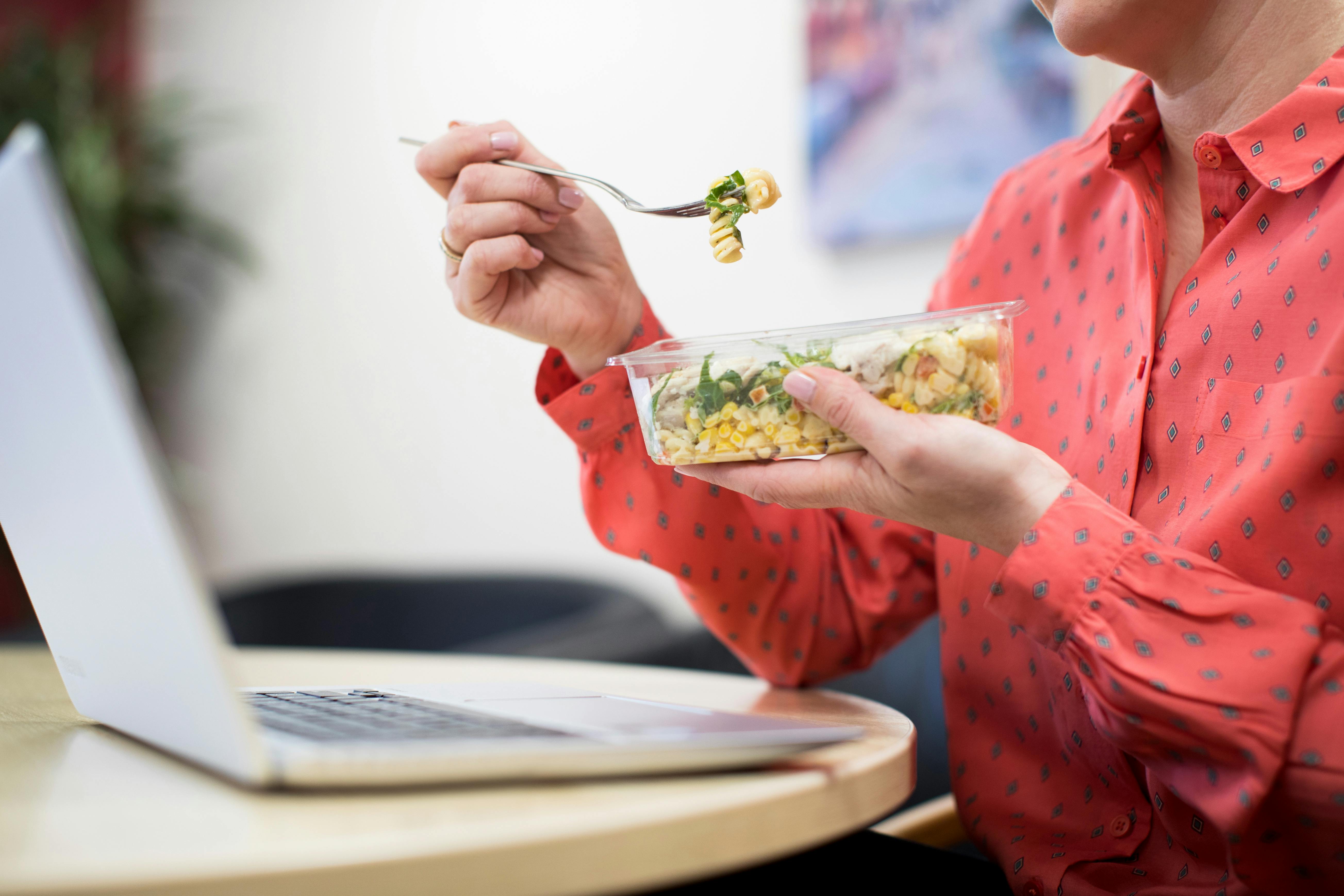 Hvis du spiser for hurtigt, påvirker det din fordøjelse. Kvinde spiser pastasalat og kigger på computer