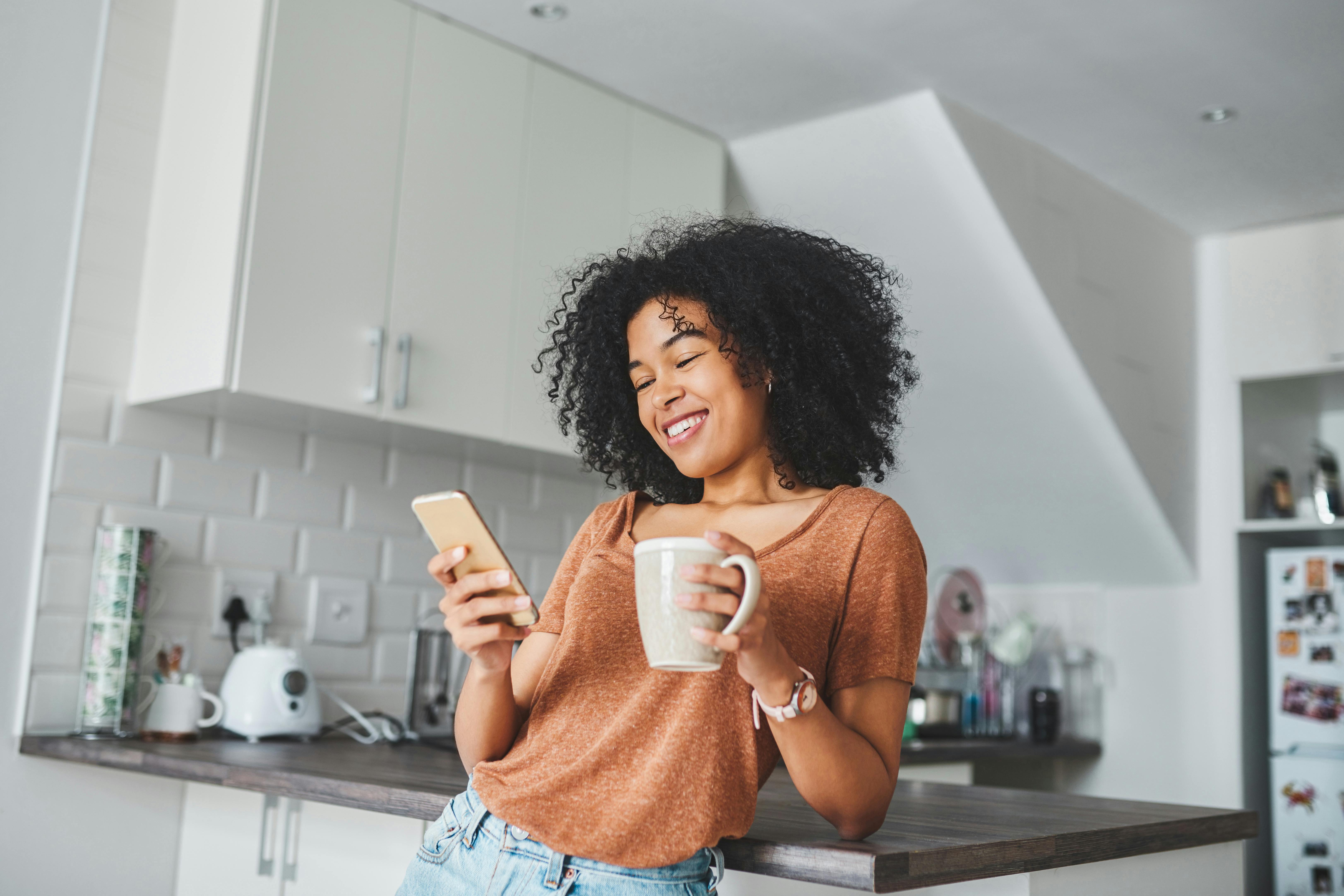 Kvinde ser på sin telefon og drikker kaffe i køkkenet