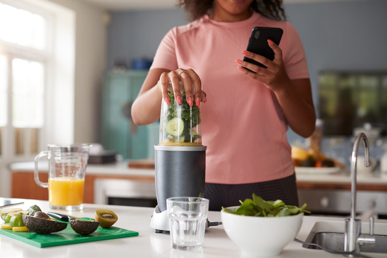 Håll koll på ditt proteinintag – särskilt om du tränar. Kvinna mixar grönsakssmoothie och kollar på mobilen