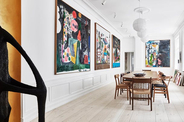 De grote, kleurrijke schilderijen van Tal R hebben een perfecte plek gekregen in de grote eetkamer. Maar kijk goed aan de eettafel, alle stoelen zijn anders, allemaal klassiekers. Grote eetkamer met houten eettafel en Deense stoelen