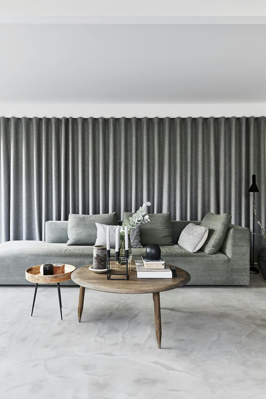 GEDEKTE KLEUREN domineren de woonkamer. Salie groene sofa, met ronde salontafels op betonvloer.