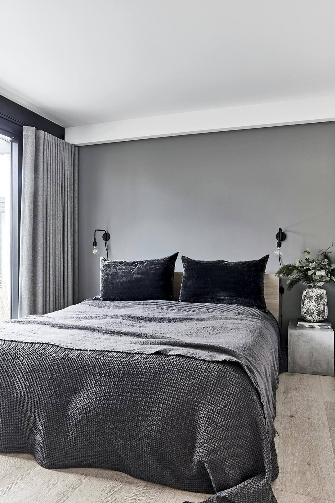 IN DE SLAAPKAMER is grijs de hoofdmoot, met gordijnen van Fischer Gardiner en beddengoed van Atelier du Noir. Het betonblok was eigenlijk voor de tuin bestemd, maar doet nu fraai dienst als nachtkastje. Het bed is van Ikea en de wandlampen zijn van Menu. Slaapkamer met grijze tinten en bed met grijze sprei.