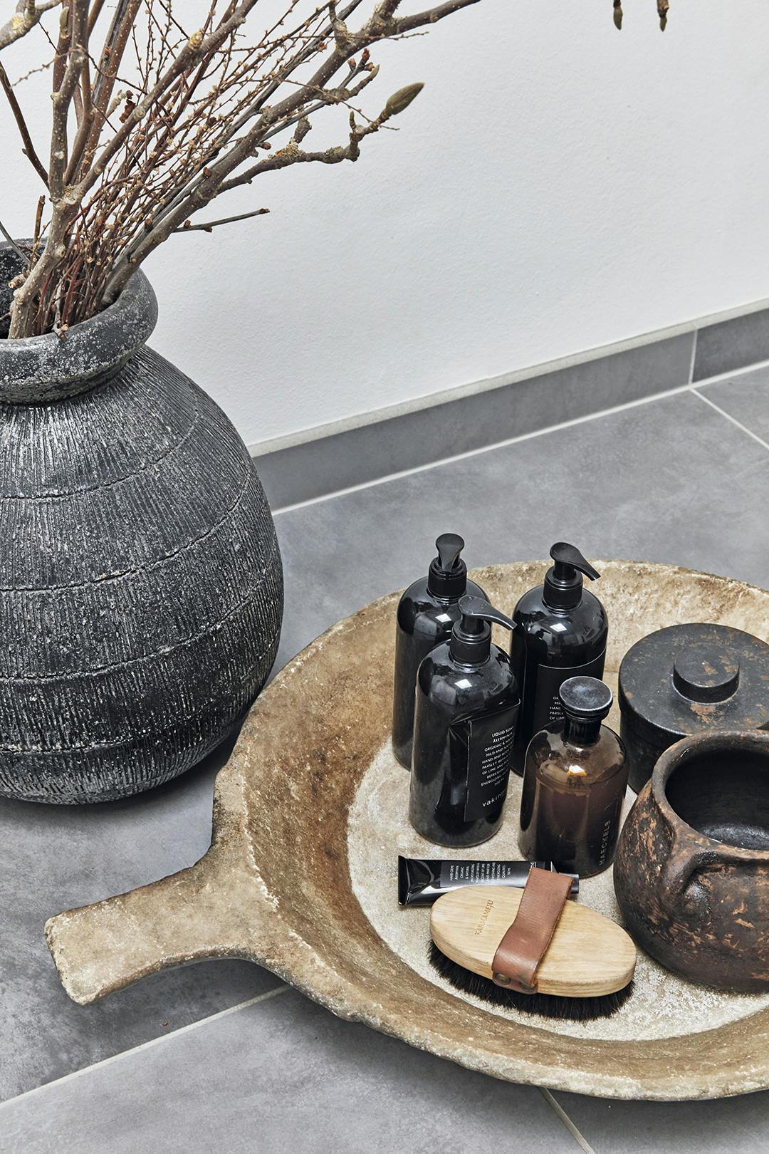 EEN KERAMIEKEN VLOERVAAS en een marmeren schaal van Atelier du Noir breken de grijze lijnen in de badkamer organisch. Badkamervloer met robuuste schaal en vaas.