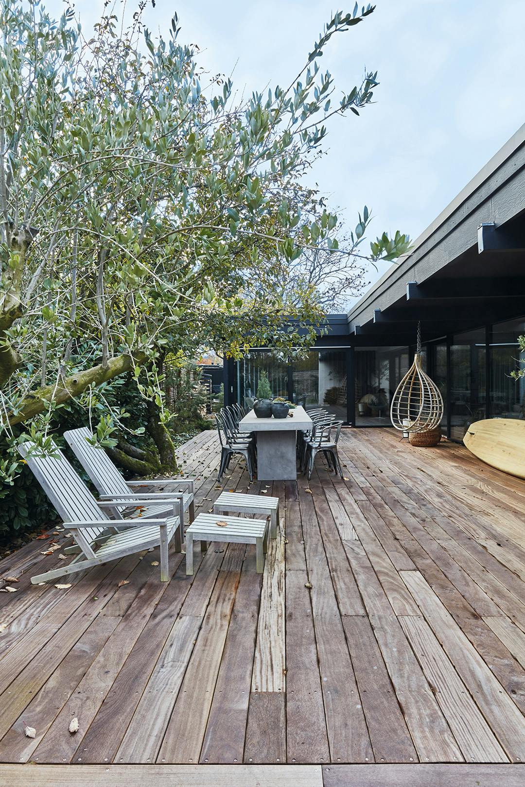 HET LANGE, HOUTEN TERRAS is op diverse manieren te gebruiken. Het tuinmeubilair van teak is van Skagerak en de hangstoel van Broste Copenhagen. Van twee betonnen tafels van Ilva is één lange tafel gemaakt. Patio tuin met houten vloer, betonnen tafel en hangstoel.
