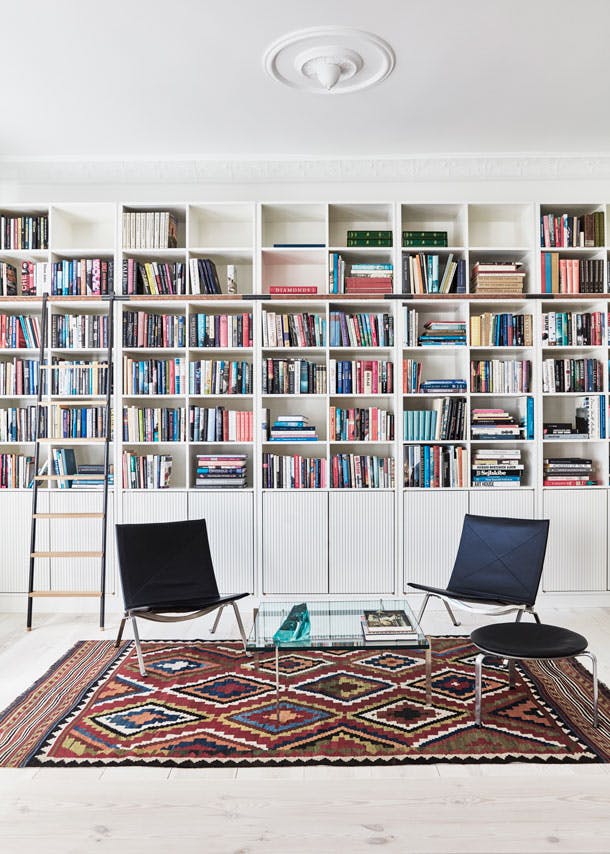De boekenast is op maat gemaakt en ervoor staan de klassieke stoelen en tafel van Poul Kjærholm. hoge witte boekenkast met ladder in scandinavische woonkamer