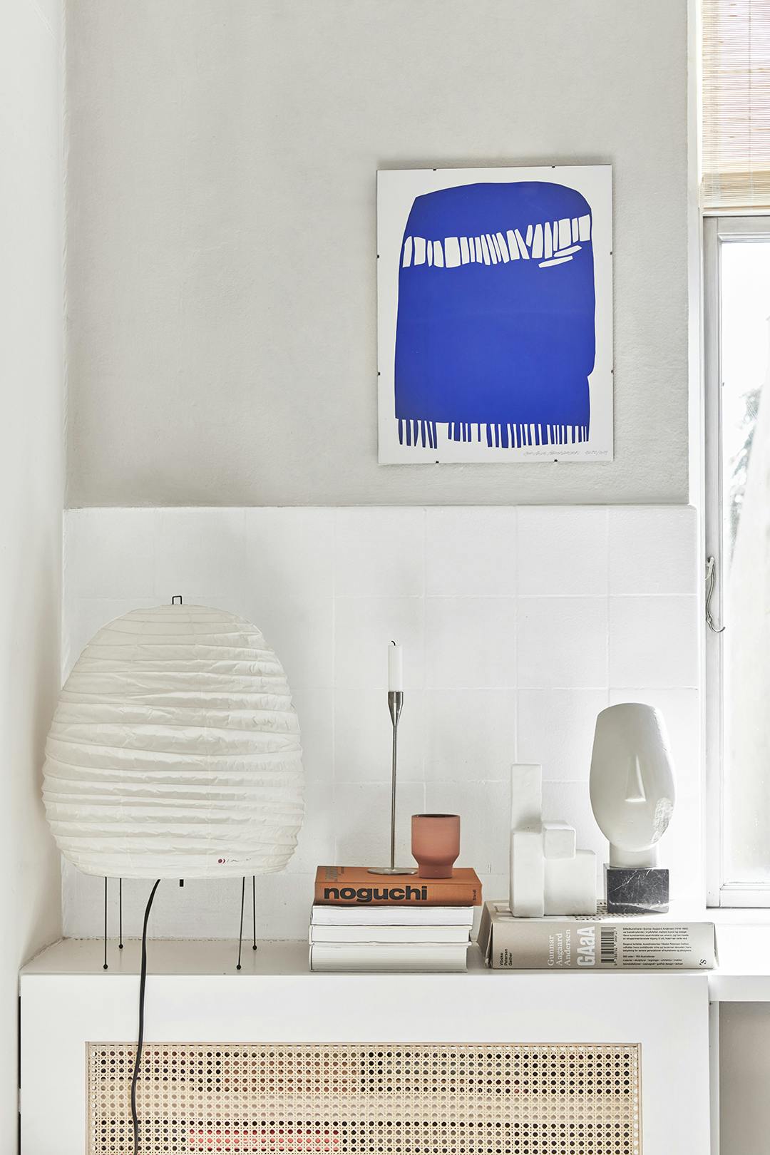 Een kleine lamp van Isamu Noguchi. Op de boeken staat werk van Atelier Cph en een vintage kandelaar van Piet Hein. Het kobaltblauwe werk is van Leise Abrahamsen. Radiatorombouw, met Noguchi lamp en Piet Hein kandelaar.