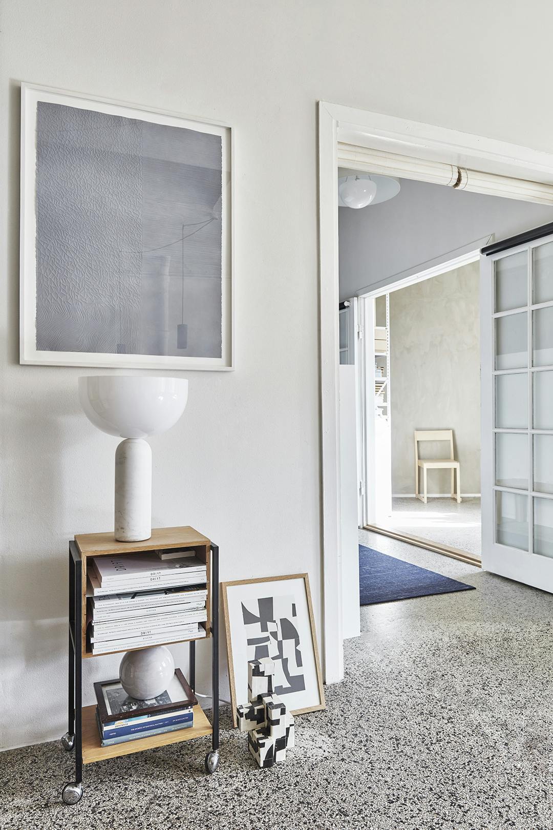 Je kunt dwars door het appartement heen kijken. Sara’s eigen ontwerpen worden geflankeerd door de sculpturale lamp van New Works op een vintage houten meubel. Erboven hangt een werk van Anders Bülow. Atelier Cph, New Works lamp en terrazzovloer.