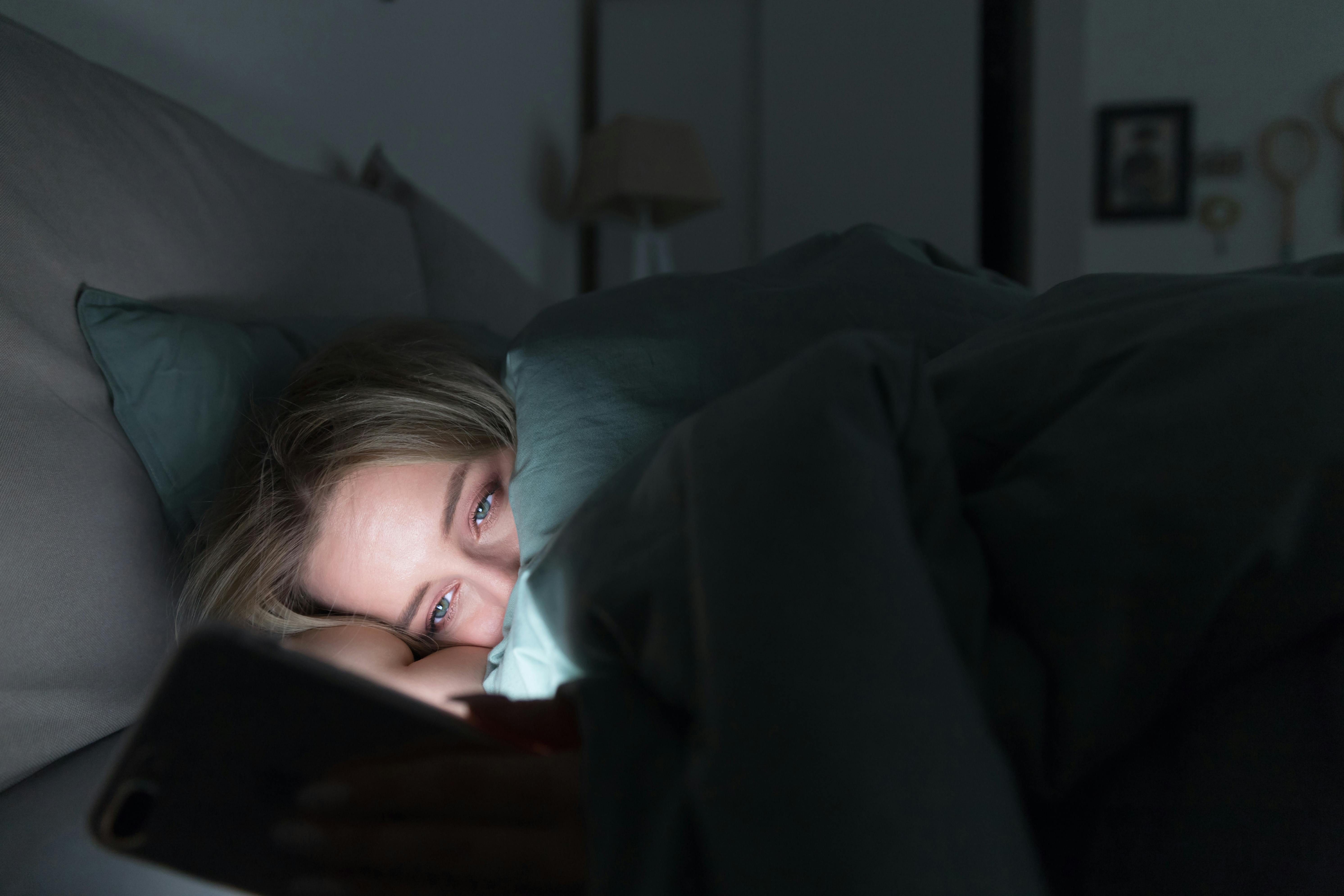 Genkender du den her situation? Der kan være flere forskellige årsager til, at du kæmper med dårlig nattesøvn. Kvinde kigger på mobil i sengen